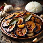 Filipino spicy eggplant adobo served in a round platter with steam, with white rice in a separate bowl in the background, with bulbs and cloves of garlic, and whole red chillies surrounding the dish