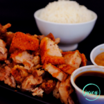 Crispy Filipino lechon kawali serve on a black rectangular platter, with steamed jasmine rice in the background, with lechon sauce, and spiced vinegar