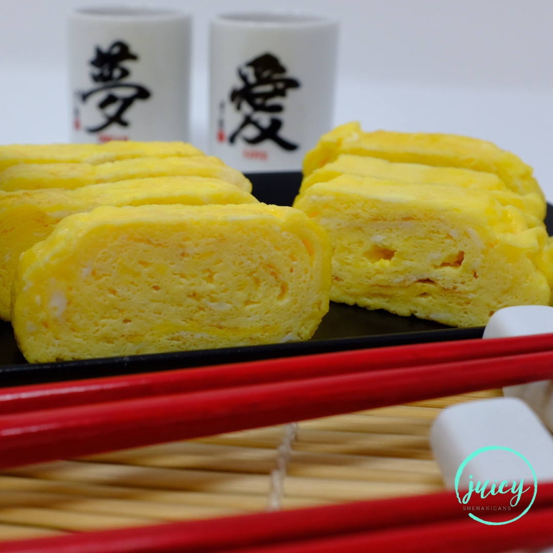 Tamagoyaki, Japanese rolled omelette, served on a black rectangular platter, displaying it's beautiful rolled layers. Red chopsticks and small tea cups in the background.