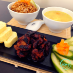 Homemade Pork Tocino, also known as Filipino cured pork, served on a black platter together with tamagoyaki and pickled vegetables, also served with a sides of rice and soup pictured in the background.